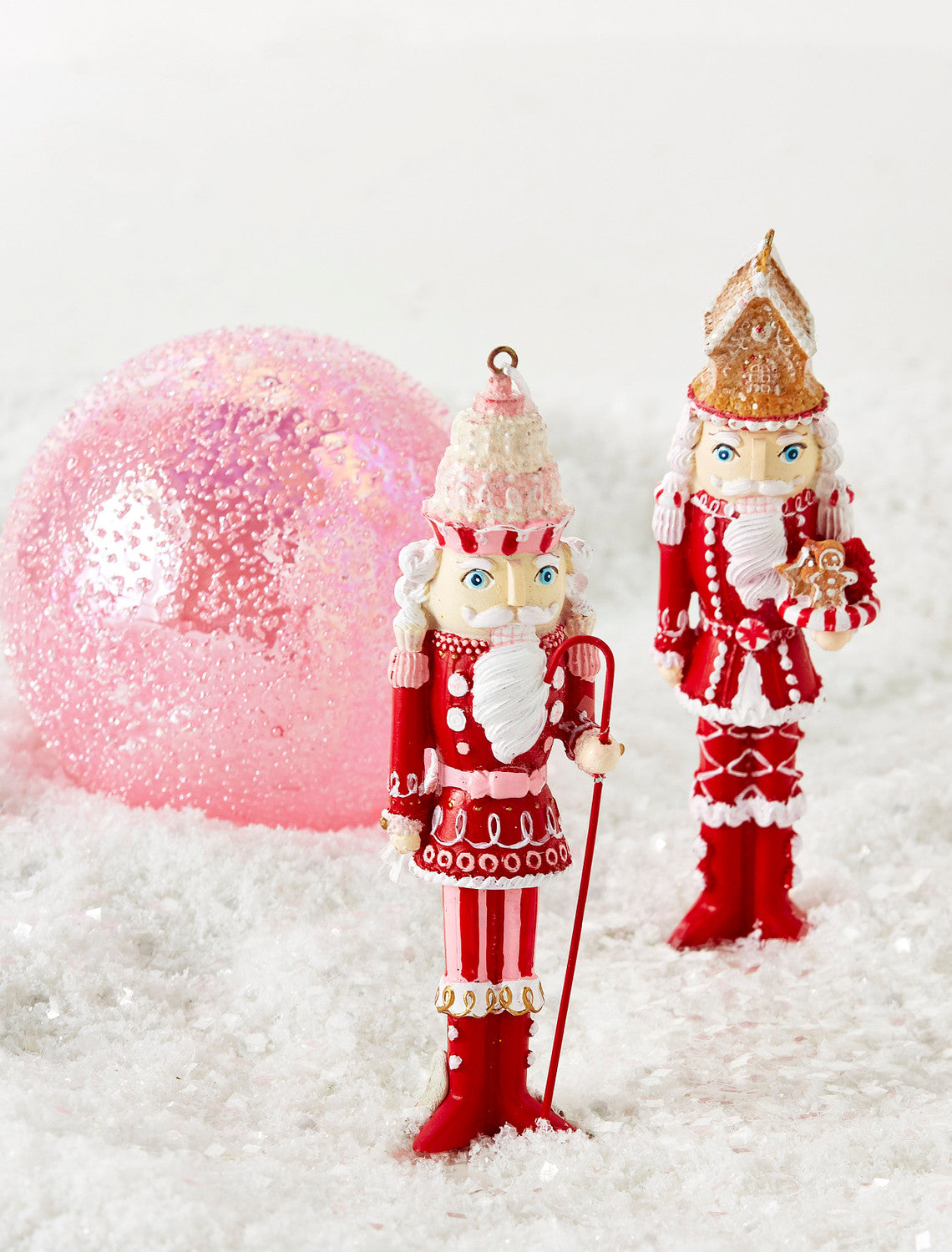 2 styles of Gingerbread Nutcracker Ornaments arranged on a table with artificial snow and a pink ornament.