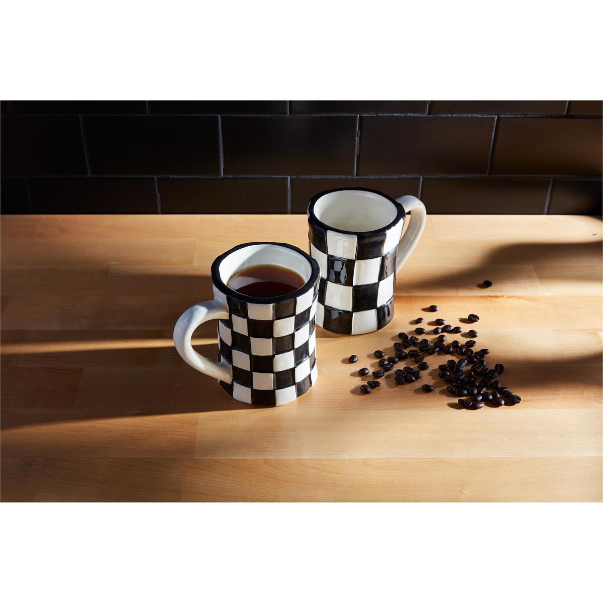 2 black and white checked mugs, one filled with coffee, on a wooden table with coffee beans scattered about.