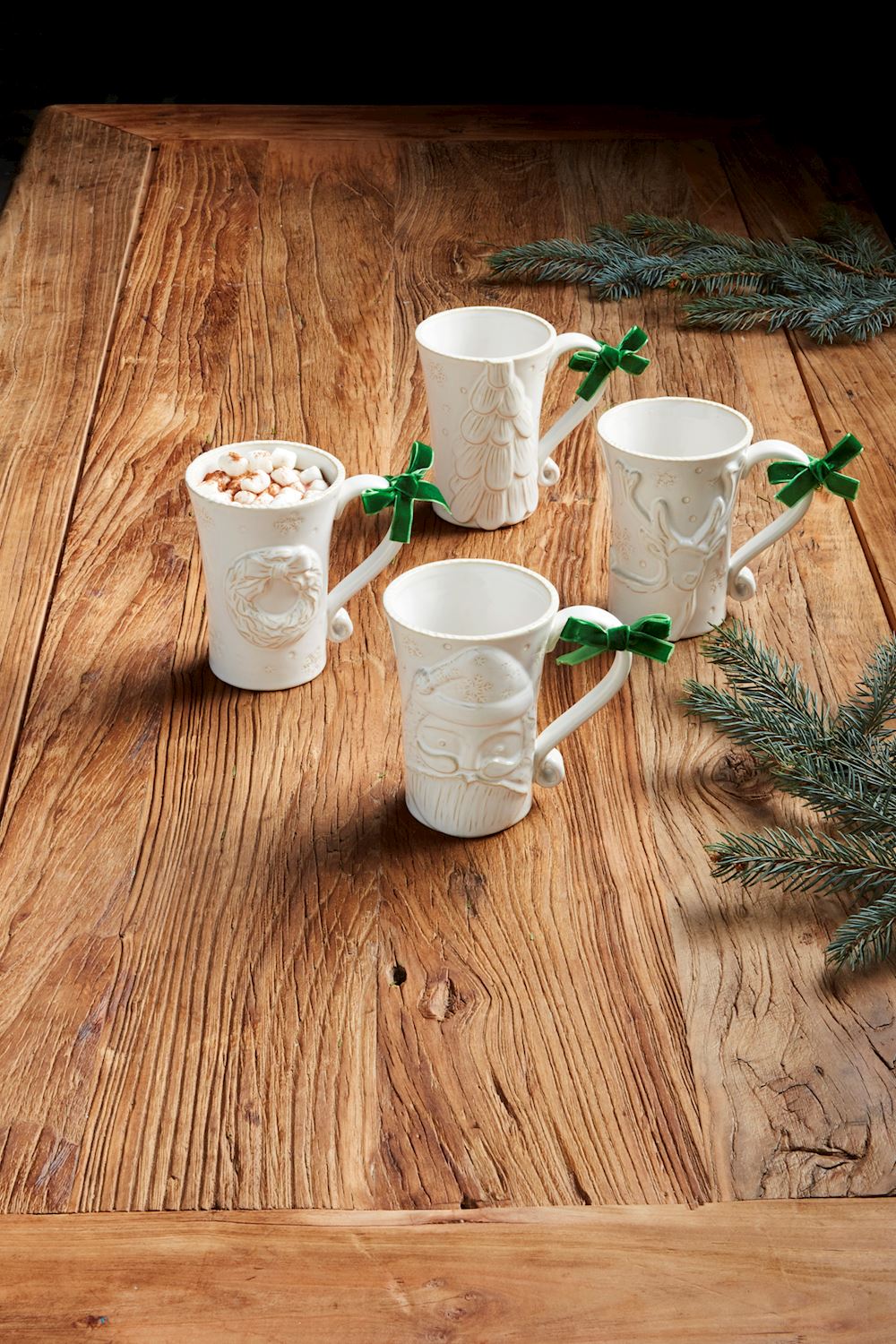 4 styles of White Christmas Mugs arranged on a wooden table with greenery.