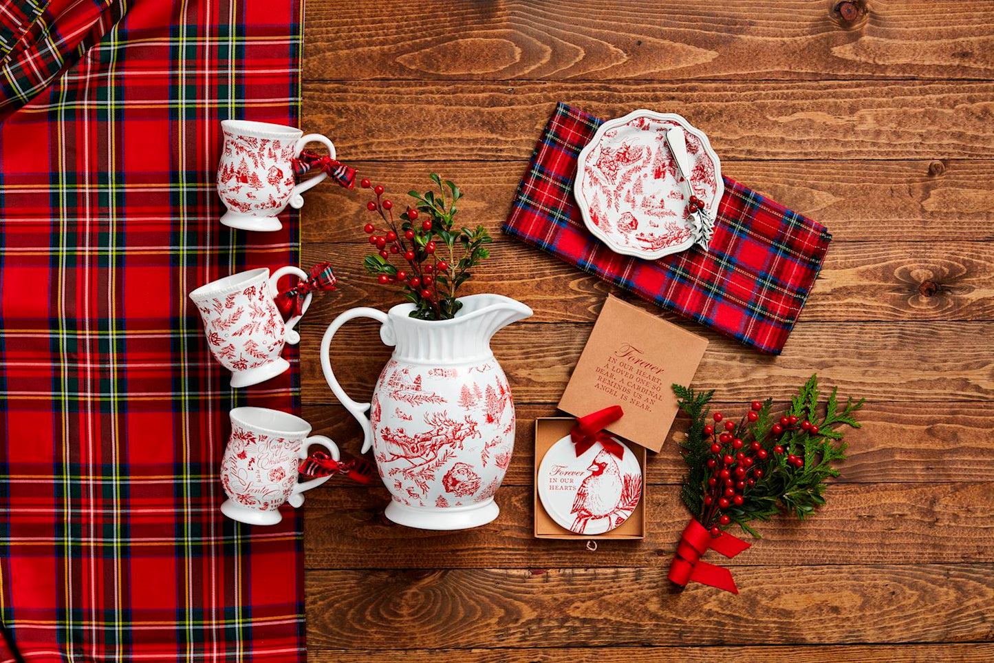 top view of a wooden table arranged with toile mugs, a pitcher, and plate with a tartan table runner and sprigs of greenery.