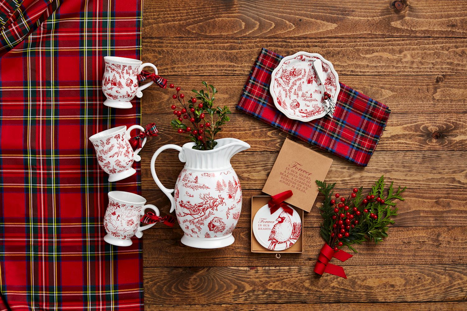 top view of a wooden table arranged with toile mugs, a pitcher, and plate with a tartan table runner and sprigs of greenery.