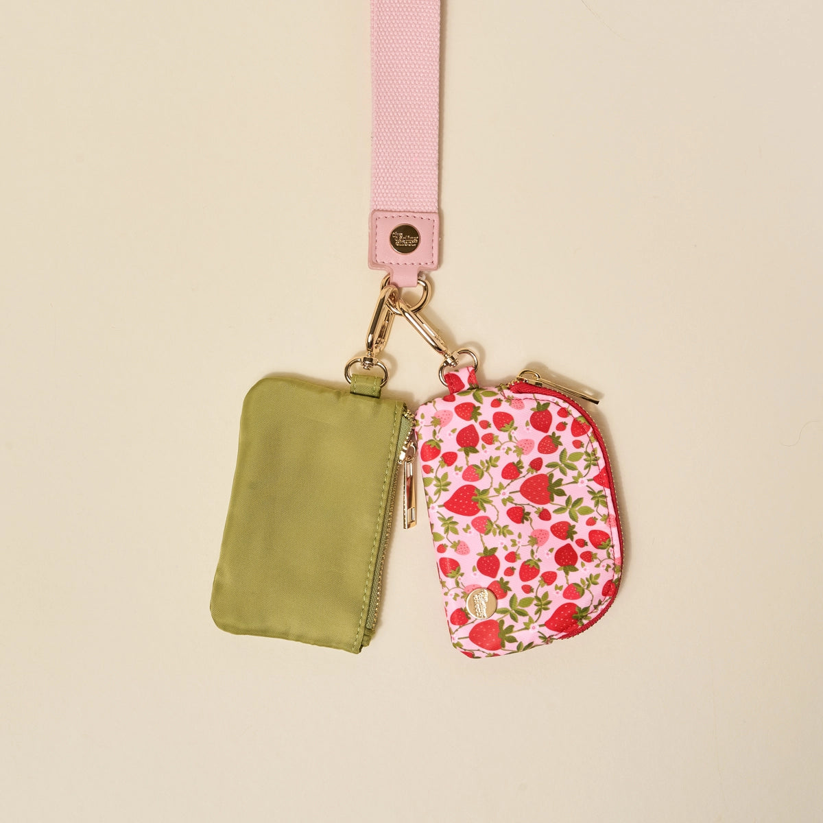 Two small pouches, one green and one with a strawberry pattern attached to a light pink strap, on a beige background.