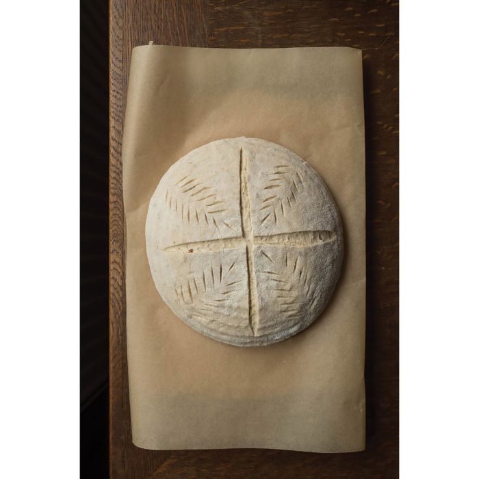 Round loaf of bread with hatch mark cuts on a brown paper on a wooden surface