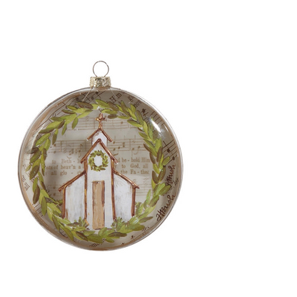 A round glass Christmas ornament featuring a painted white church with a green wreath, surrounded by a green laurel wreath and a background of vintage sheet music 