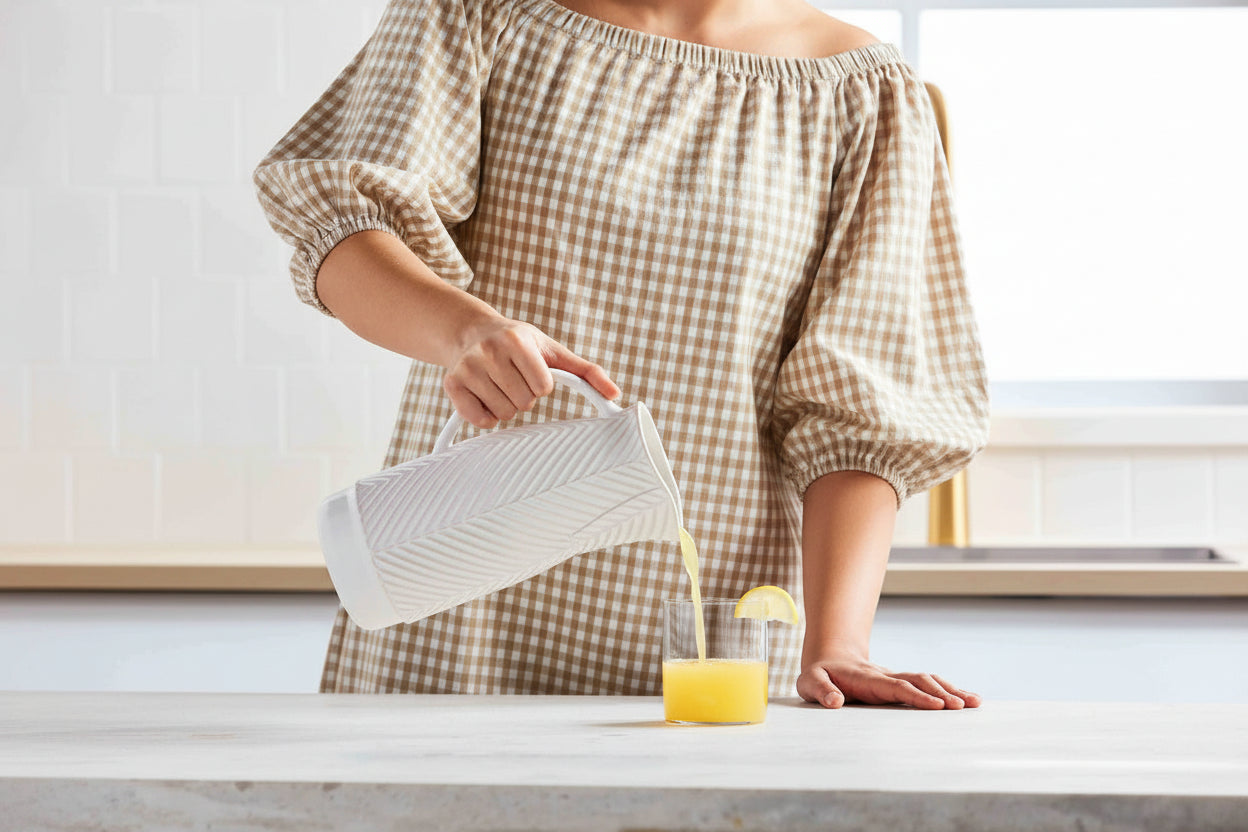 Person in a brown gingham outfit pouring a drink from a white pitcher into a glass on a light-colored surface.