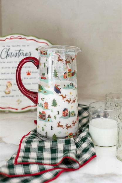 christmas village pitcher filled with milk and displayed on a counter with a green checked towel, platter, and cup of milk