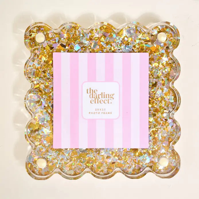 A scalloped clear photo frame filled with gold and iridescent glitter, with a pink striped insert card reading 'the darling effect' in the center, shown on a light beige background.