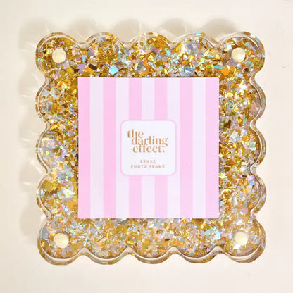 A scalloped clear photo frame filled with gold and iridescent glitter, with a pink striped insert card reading 'the darling effect' in the center, shown on a light beige background.