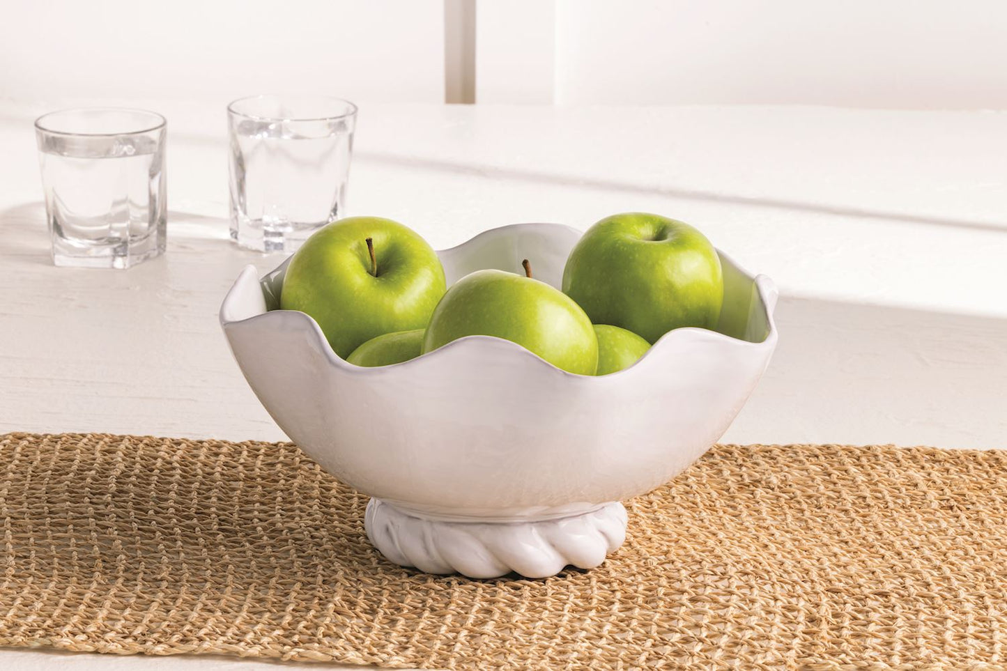 white ceramic bowl with a twisted base filled with green apples and set on a jute table runner