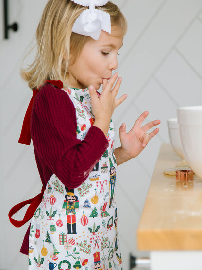 child wearing nutcracker apron, licking their finger, and standing at a counter filled with mixing bowls and cookie cutters.