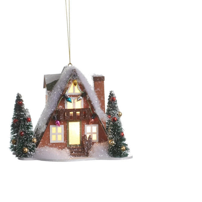 A Christmas ornament shaped like a snow-covered cabin with warm glowing windows, decorated with small Christmas trees on both sides, hanging from a thin gold string against a plain white background.