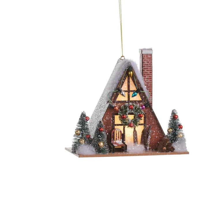 A decorative Christmas ornament shaped like a small snow-covered house with a lit window, brick chimney, garlands, red ornaments, and two small decorated trees, hanging by a gold string on a white background.