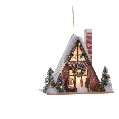A decorative Christmas ornament shaped like a small snow-covered house with a lit window, brick chimney, garlands, red ornaments, and two small decorated trees, hanging by a gold string on a white background.