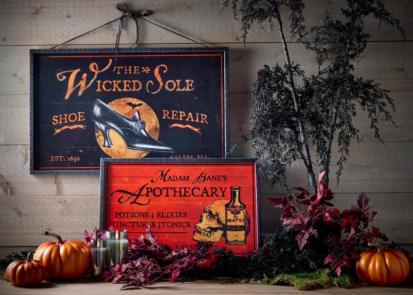 Decorative signs with Halloween-themed text and images on a rustic wooden background with fall foliage and pumpkins on a table below them