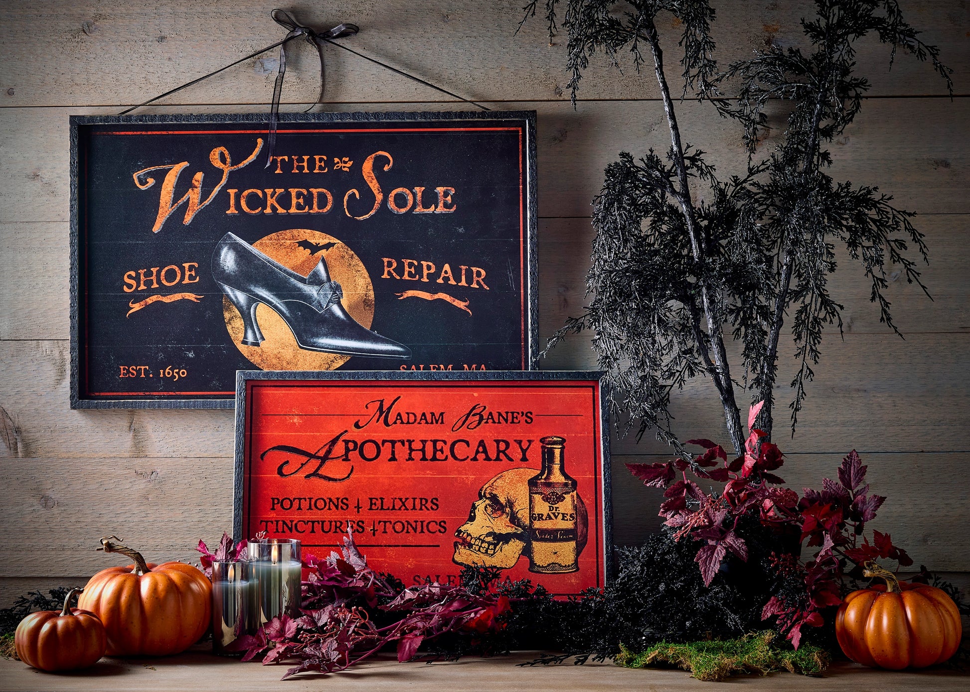 Decorative signs with Halloween-themed text and images on a rustic wooden background with fall foliage and pumpkins on a table below them