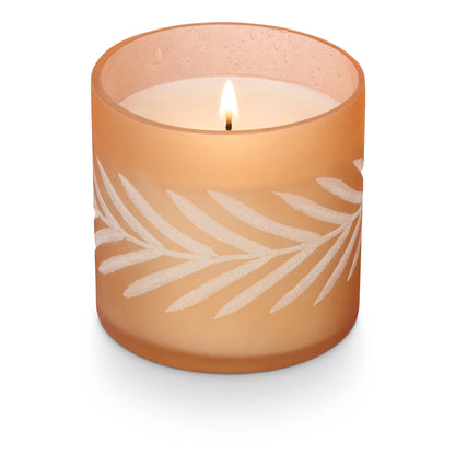 A soy wax candle in a frosted glass jar with a leaf pattern, with a lit flame.