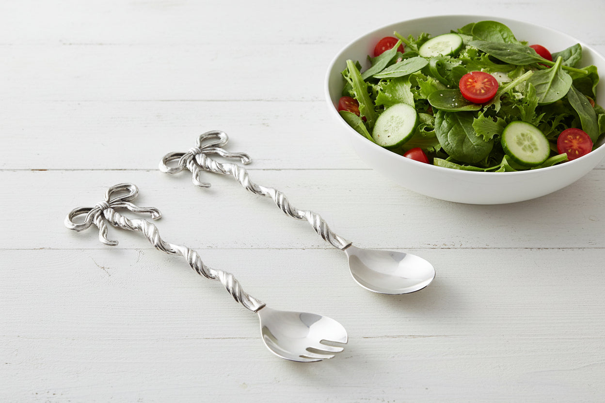 metal serving utensils with bow handles set on a table with a bowl of salad