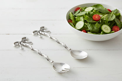 metal serving utensils with bow handles set on a table with a bowl of salad