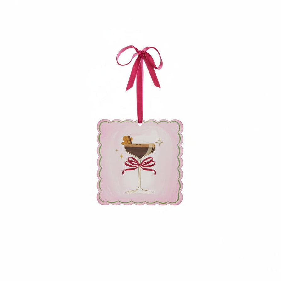 A square pink ornament with scalloped edges, featuring an illustration of a martini glass with a red bow on its stem and a gingerbread man garnish