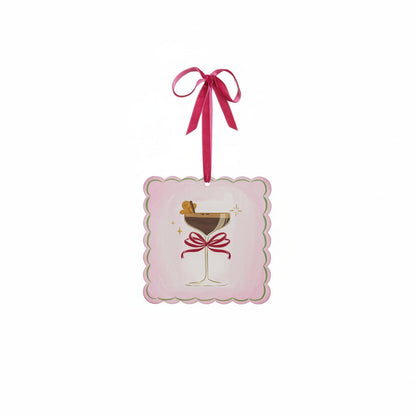 A square pink ornament with scalloped edges, featuring an illustration of a martini glass with a red bow on its stem and a gingerbread man garnish