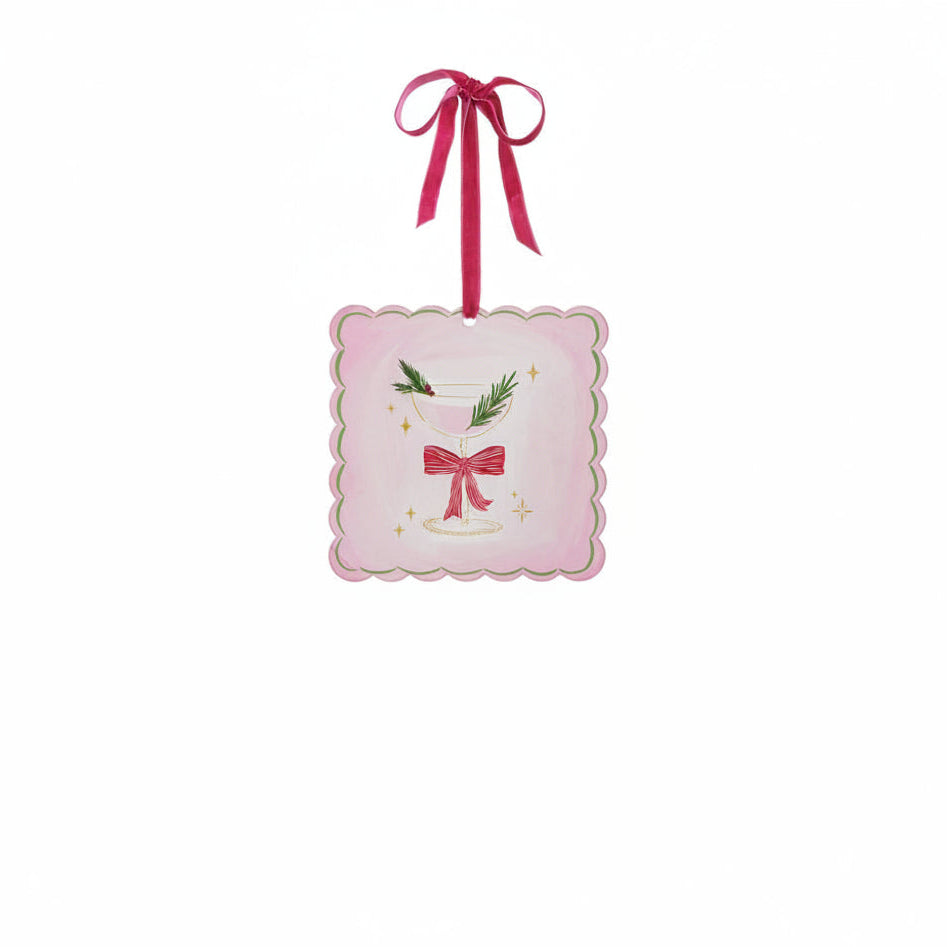 A square pink ornament with scalloped edges, featuring an illustration of a martini glass with a red bow on its stem and a sprig of greenery with red berries as a garnish