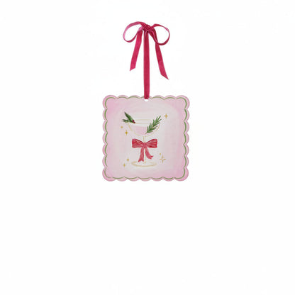 A square pink ornament with scalloped edges, featuring an illustration of a martini glass with a red bow on its stem and a sprig of greenery with red berries as a garnish
