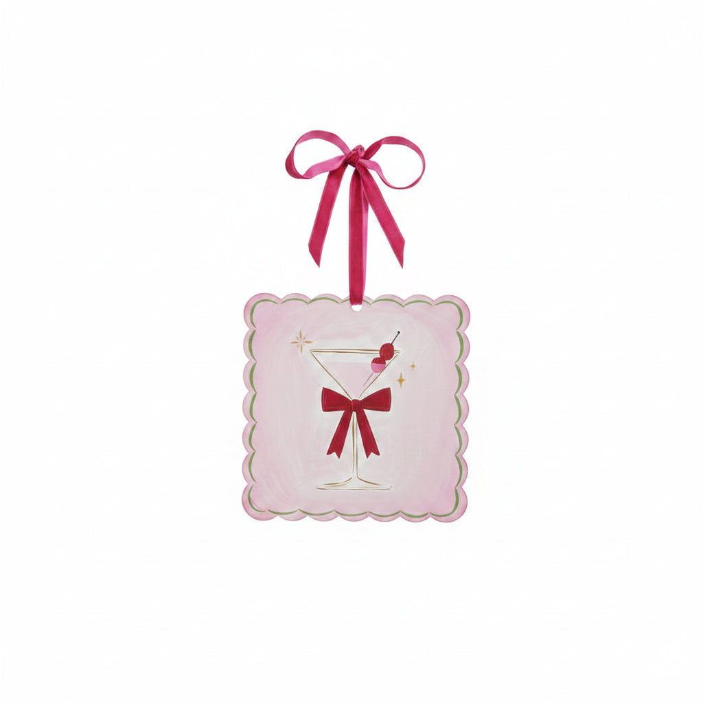 A square pink ornament with scalloped edges, featuring an illustration of a martini glass with a red bow on its stem and olive garnishes
