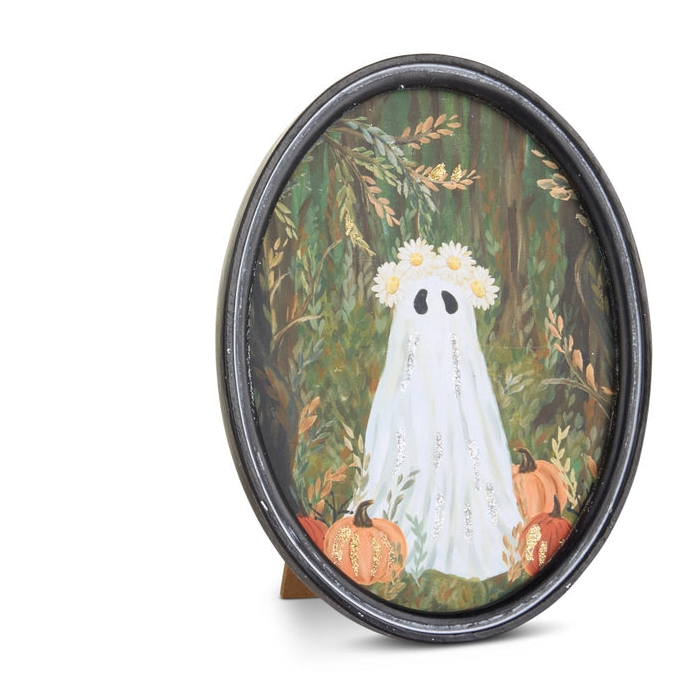 An oval framed art print featuring a whimsical painting of a white ghost with a flower crown standing in a forest with pumpkins, displayed on a small wooden easel against a white background.
