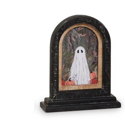 A Halloween-themed decoration in a  black arched frame with an illustration of a white ghost surrounded by pumpkins in an autumn forest setting.