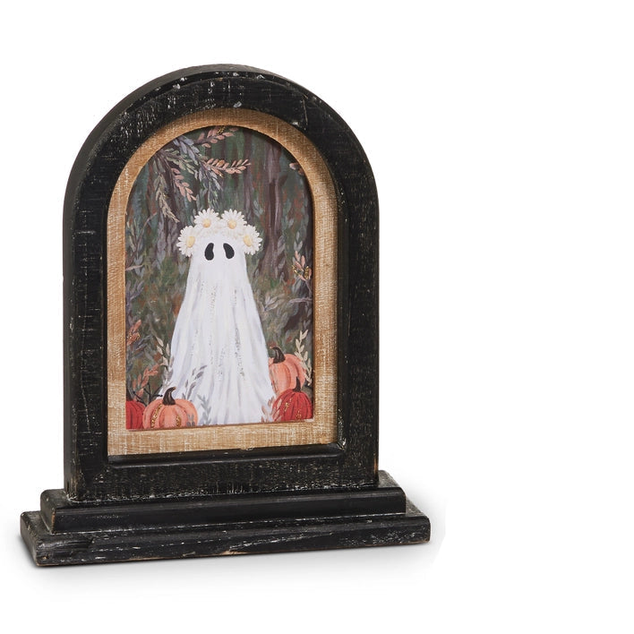 A Halloween-themed tabletop decoration featuring a distressed black arched frame with a painted illustration of a white ghost wearing daisies around the head surrounded by pumpkins on a light wood background.