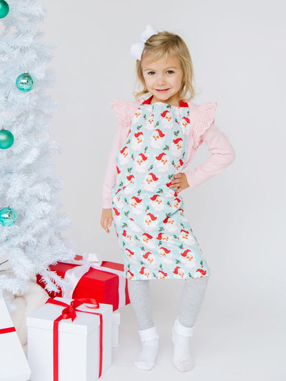 child wearing santa apron posing next to a tree with packages under it.