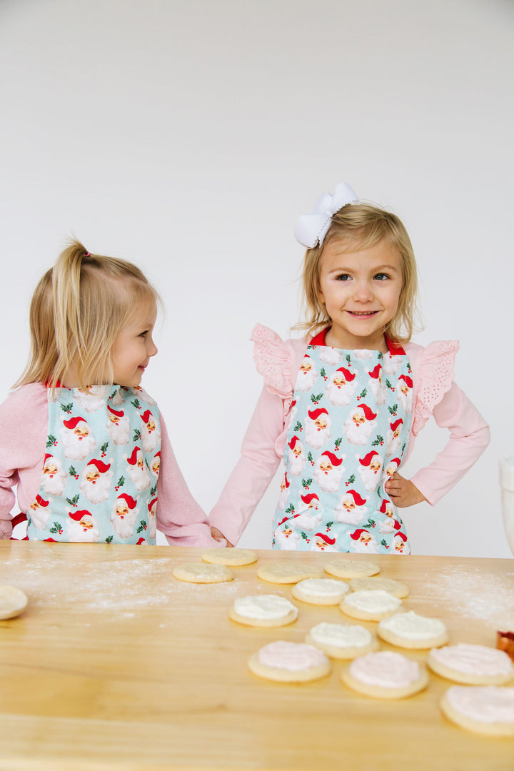 2 children wearing santa aprons standing at a counter filled with cookies.