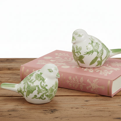 Two ceramic birds with green floral patterns on a wooden table, one bird in set on a pink book