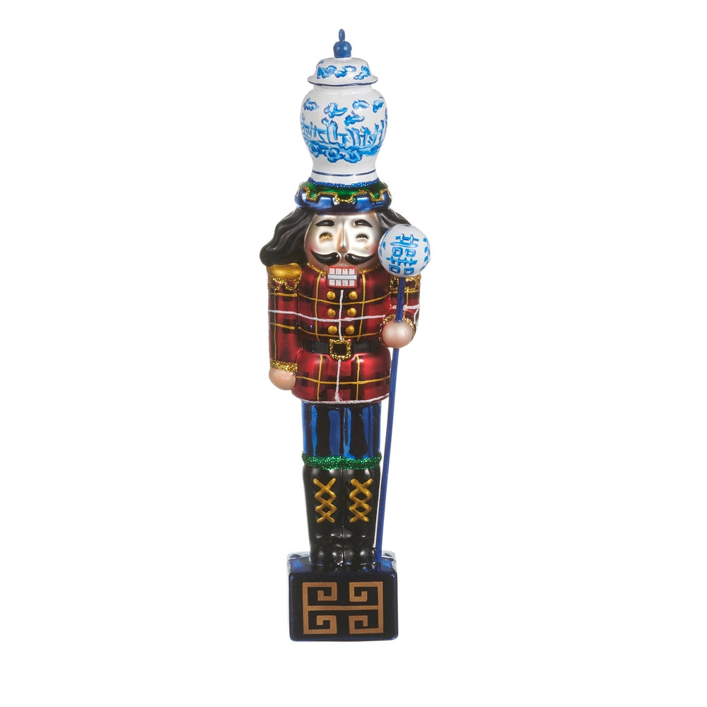 glass nutcracker ornament with wearing red plaid coat and bleu pants with a blue and white ginger jar on its head and holding a staff