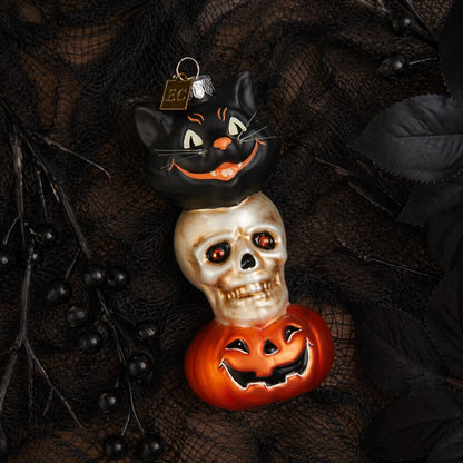 A stacked Halloween glass ornament featuring a black cat on top of a skull, which is on top of a carved orange pumpkin, styled on a dark black lace fabric background.
