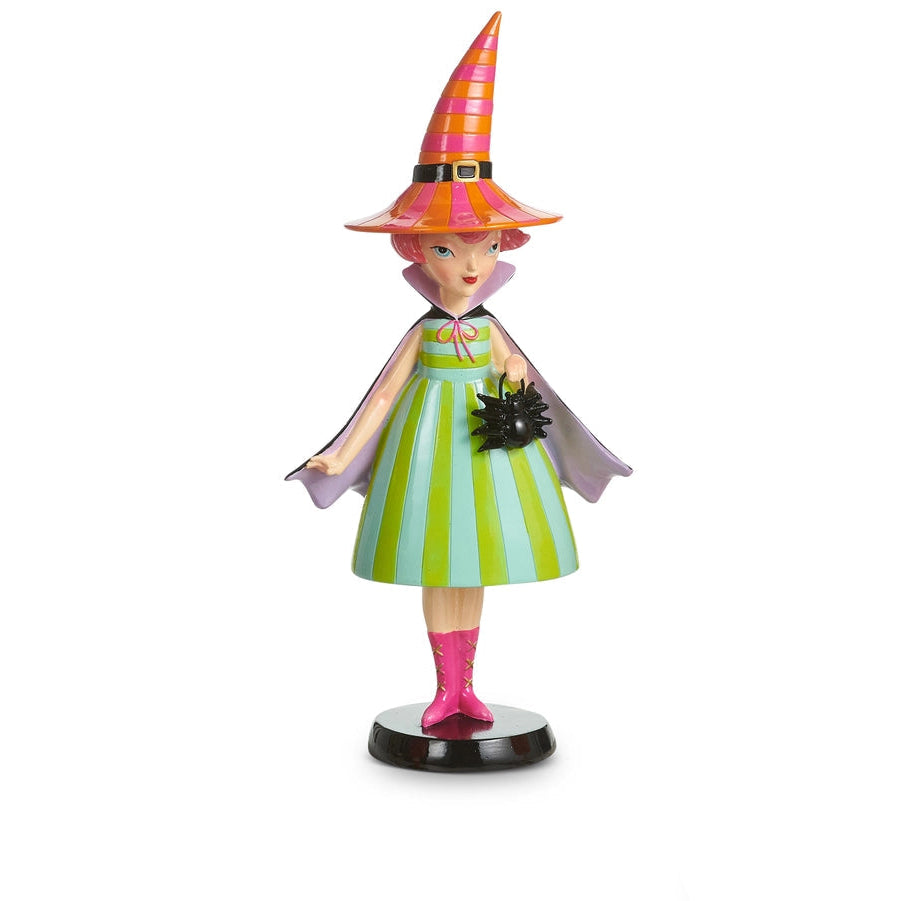 witch figurine wearing a green and blue striped dress, black cape, and orange and pink striped witches hat. she is holding a spider shaped handbag