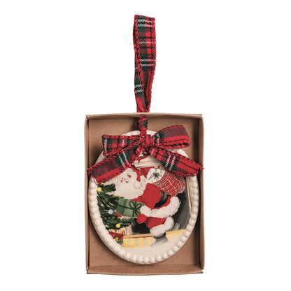 A oval Christmas ornament featuring Santa Claus coming out of a fireplace next to a tree, presented in its brown gift box with a red plaid ribbon bow and hanging loop.