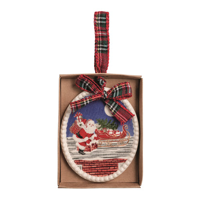 A Christmas ornament in its brown cardboard box, featuring an oval design with Santa Claus next to his sleigh on a rooftop against a blue night sky, accented with a red and green plaid ribbon bow and hanging loop.