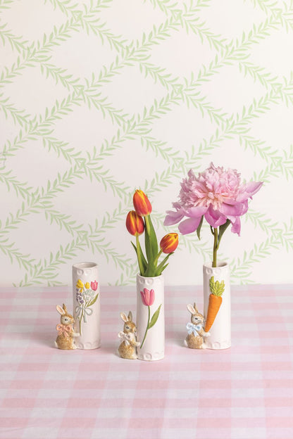 Three decorative vases with floral designs and a little brown bunny attached to the side on a pink checkered tablecloth against a light green leaf-patterned wall.