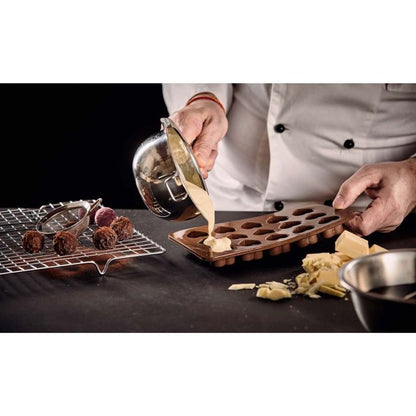 Person in a chef's coat pouring a mixture into a chocolate mold on a dark surface.