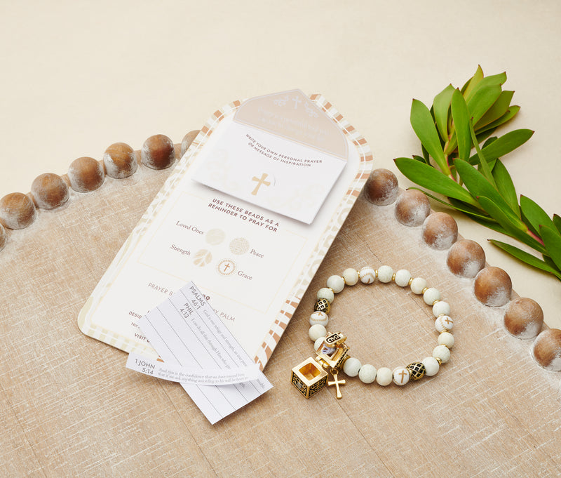 ivory beaded bracelet with gold cross and prayer box charms laying next to card packaging and prayer slips.