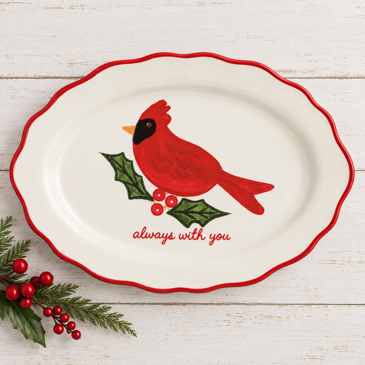 An oval white ceramic serving platter with a scalloped red rim, featuring a painted red cardinal on green holly with red berries and the words 'always with you' in red script. The platter is photographed on a white wood plank surface with holly sprigs at the lower left.