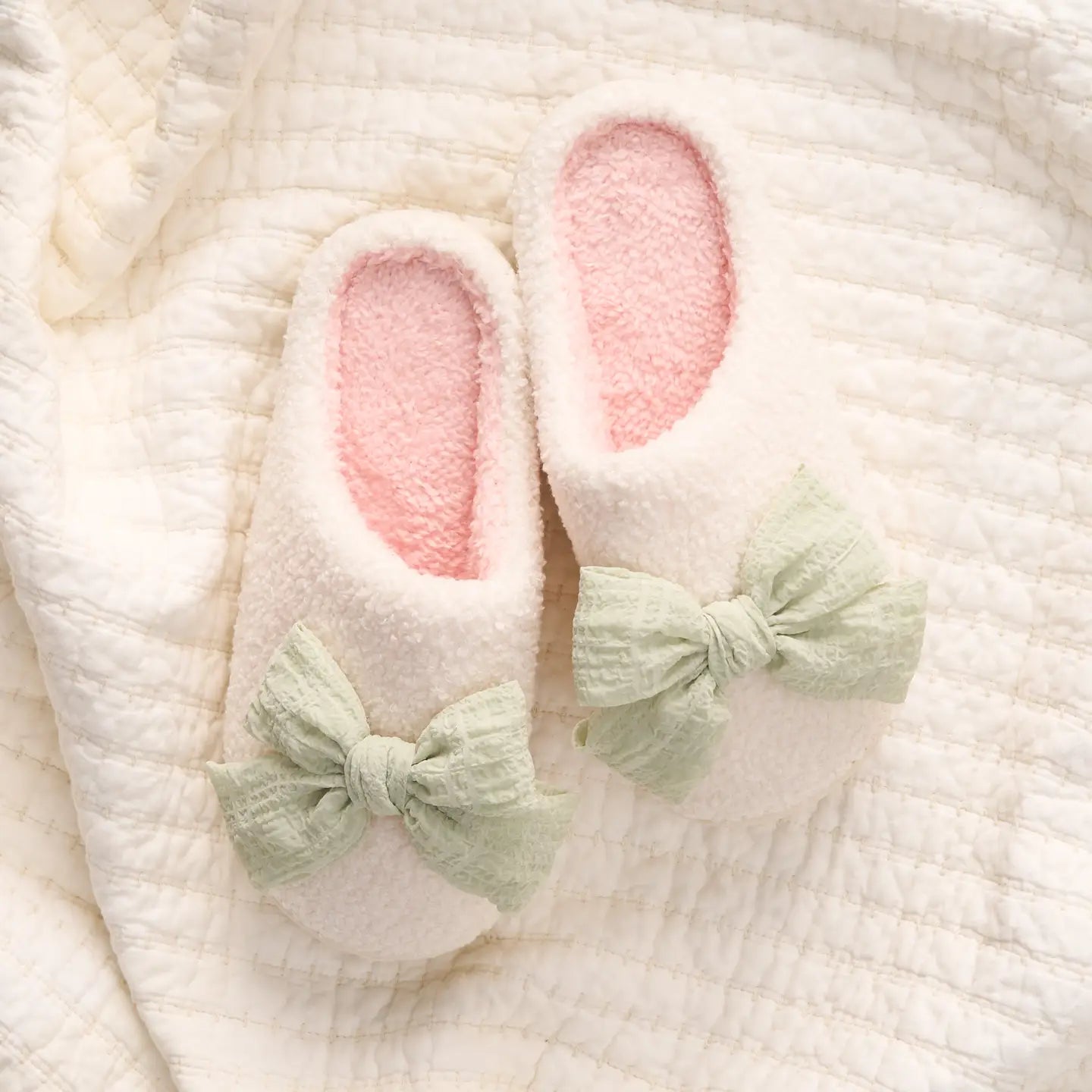 sage betty bow fuzzy slippers with a light pink insole set on a cream-colored quilt