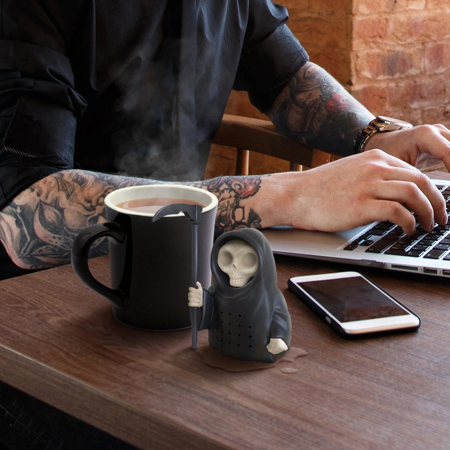 Grim Steeper Tea Infuser set on a desk next to a cup of tea with a person working of a laptop in the background.