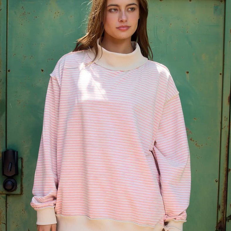 person wearing a cream mock neck sweatshirt with pink stripes on it standing in front of a green metal door