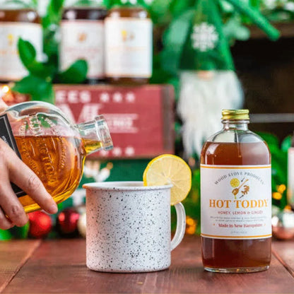 bottle of Hot Toddy Mix set on a wood table next a metal cup with a lemon on the rim and a hand pouring spirits in to it