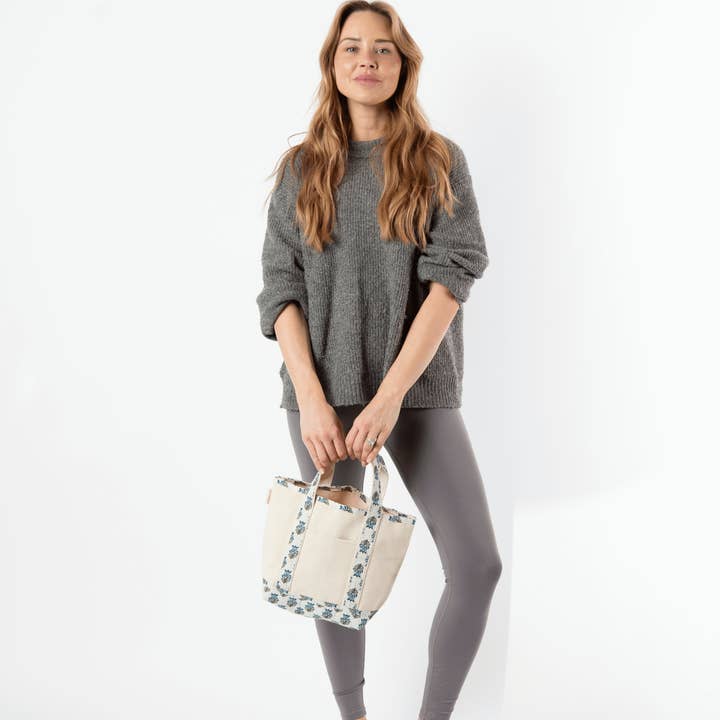 person wearing grey pants and sweater holding off white tote with aqua floral printed handles and trim