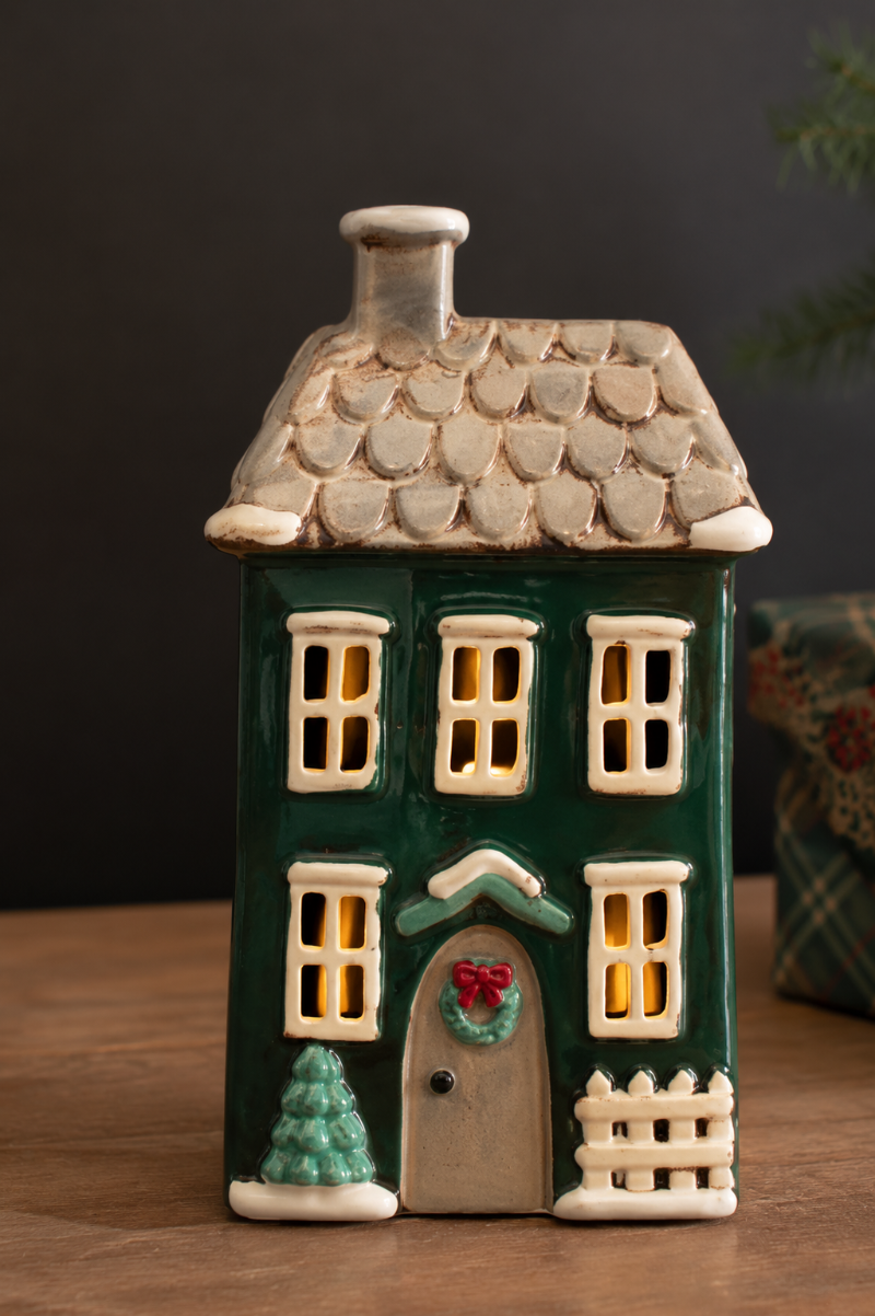 A ceramic Christmas house figurine with a dark green body, beige roof and trim, glowing yellow windows, a gray door, and small holiday accents including a wreath, Christmas tree, and picket fence, displayed on a wooden surface against a dark background.