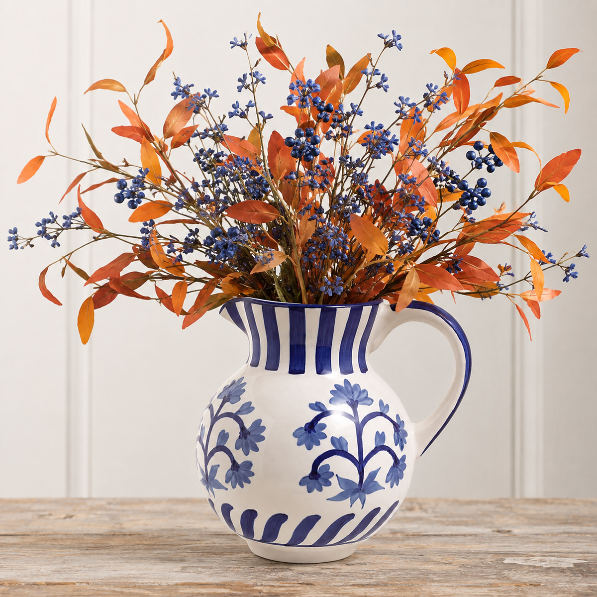 An artificial autumn floral arrangement with orange leaves and blue berries displayed in a white ceramic pitcher with blue floral patterns, set on a rustic wooden table against a white paneled wall.