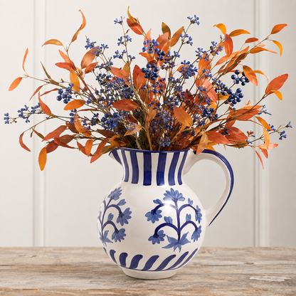 An artificial autumn floral arrangement with orange leaves and blue berries displayed in a white ceramic pitcher with blue floral patterns, set on a rustic wooden table against a white paneled wall.